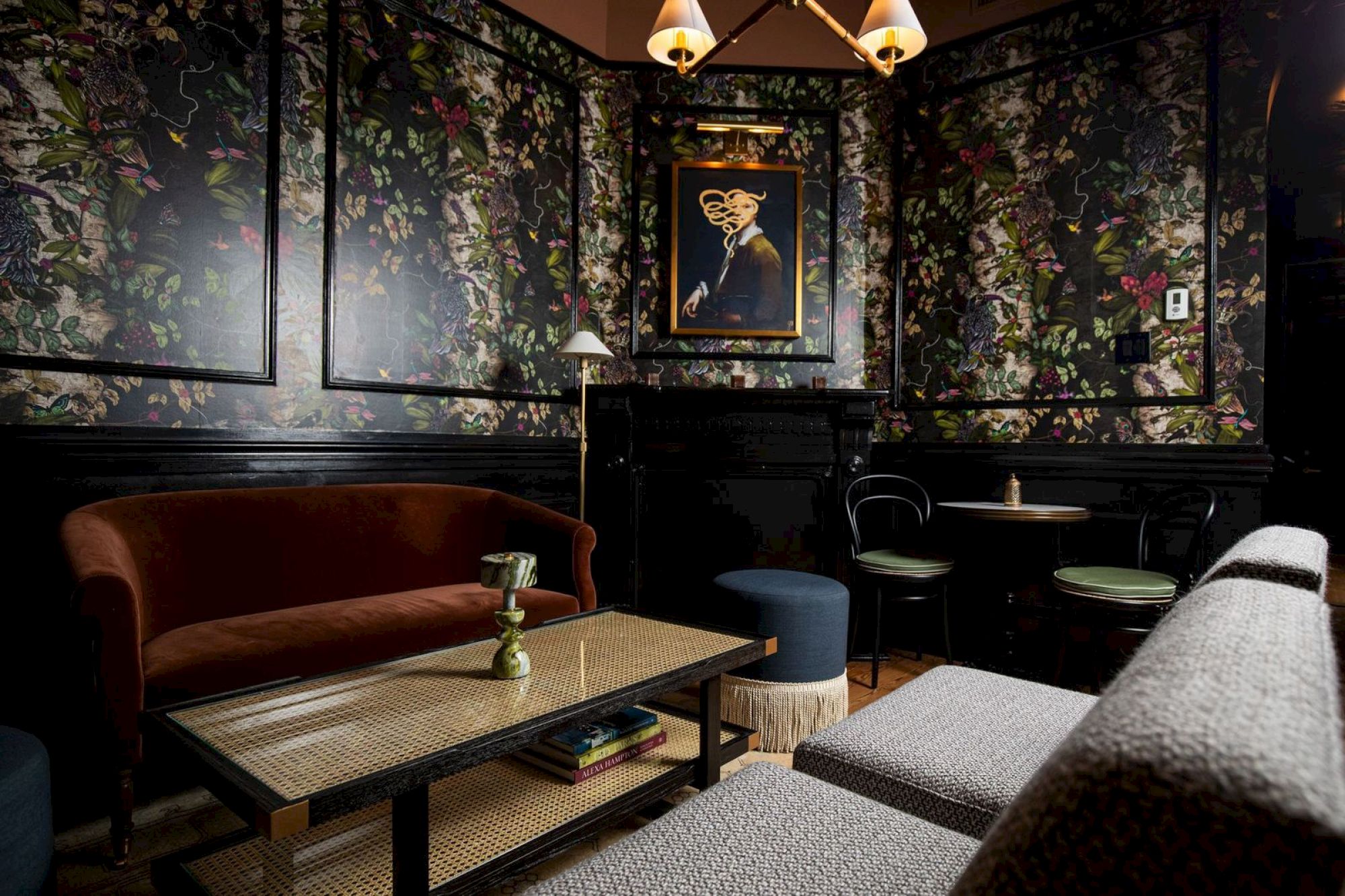 A cozy room with floral-patterned walls, a painting, a brown velvet sofa, a table with a lamp, and chairs under a chandelier.