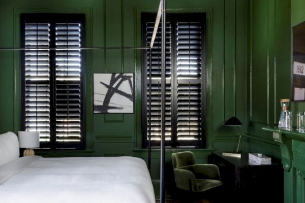 A stylish bedroom with green walls, a canopy bed, patterned rug, modern chandelier, and shuttered windows, creating a cozy ambiance.