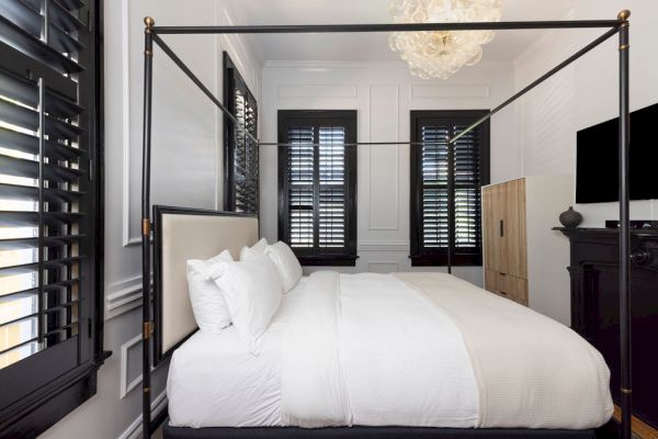 The image shows a bedroom with a four-poster bed, white bedding, black window shutters, a chandelier, and a wall-mounted TV.