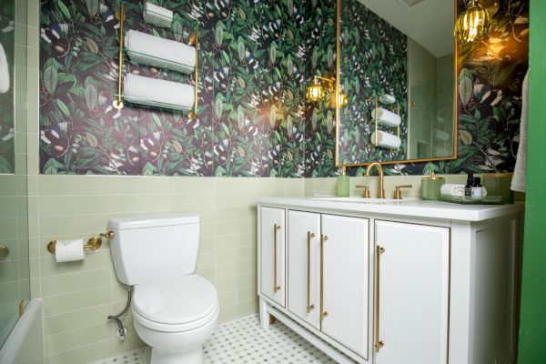 A bathroom with floral wallpaper, white cabinets, double sinks, a toilet, and gold fixtures. Towel rack and wall lights enhance decor.