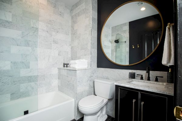 A modern bathroom features a bathtub, toilet, and vanity with a large round mirror, set against marble and black accents.