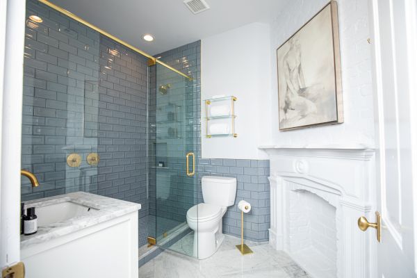 The image shows a modern bathroom with a glass shower, marble sink, toilet, and wall art, featuring gold fixtures and gray tiles.