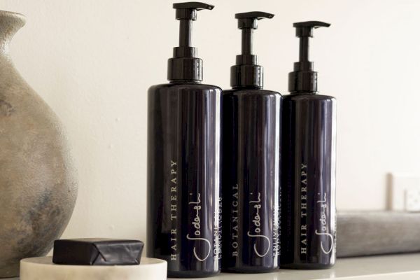 The image shows three black pump bottles labeled "Hair Therapy" on a shelf, alongside a dish with a bar of soap and a decorative vase.