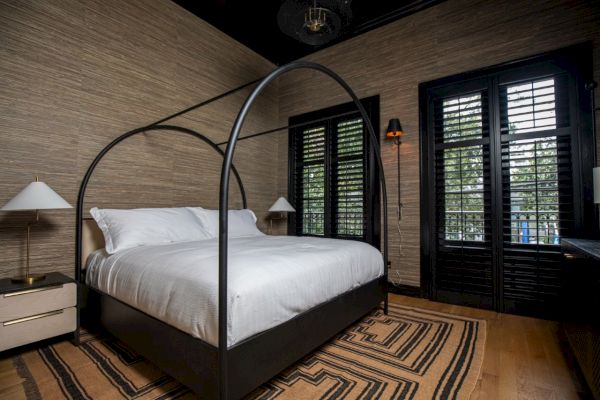 A bedroom with a canopy bed, white bedding, neutral walls, wooden shutters, lamps, and a striped rug on a wooden floor.