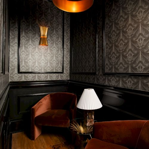 The image shows a cozy room with dark wallpaper, two brown chairs, a small table with a lamp, and warm lighting from a ceiling fixture.