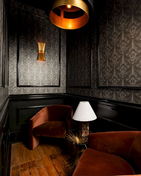 The image shows a cozy room with dark wallpaper, two brown chairs, a small table with a lamp, and warm lighting from a ceiling fixture.