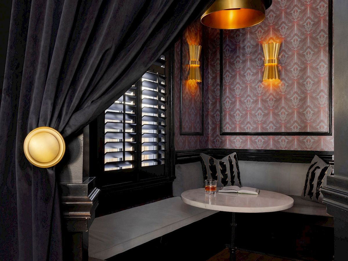 A cozy nook with dark curtains, a round table, and cushioned seating, softly lit by golden lamps and a ceiling fixture.