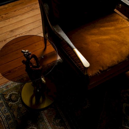 A dimly lit room featuring a wooden floor, ornate rug, leather armchair, and a small round side table with a decorative statue.