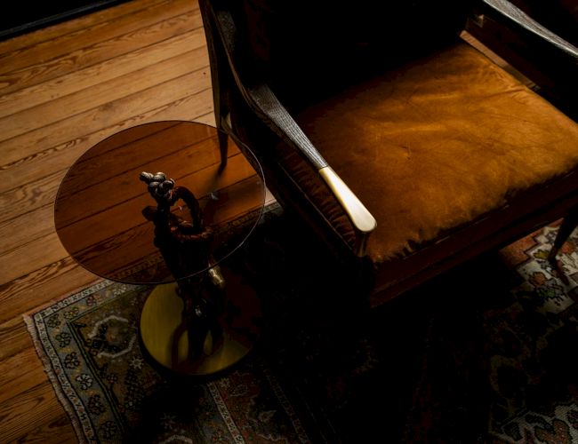 A dimly lit room featuring a wooden floor, ornate rug, leather armchair, and a small round side table with a decorative statue.