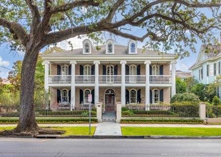 A large, elegant house with a wraparound balcony and columns, surrounded by a garden and iron fence, with a large tree in front.