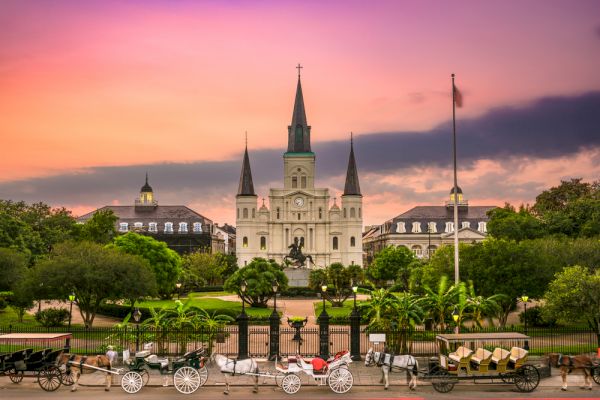 The image shows a sunset view of a historic building with spires, surrounded by greenery and horse-drawn carriages in front.