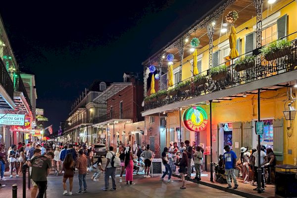 The image shows a bustling street scene at night, with people walking past vibrant buildings, neon signs, and decor, creating a lively atmosphere.