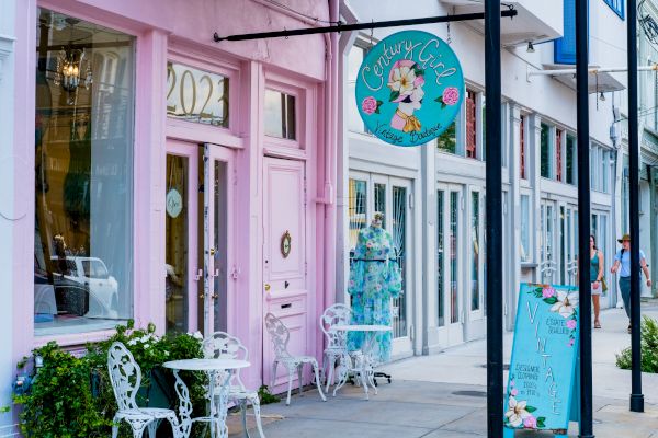 The image shows a pastel pink storefront with decorative tables and chairs outside, featuring a round sign that reads "Century Girl."