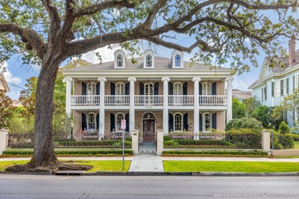 The image shows a large, elegant two-story house with white columns, symmetrical windows, and a manicured lawn, framed by a large tree.
