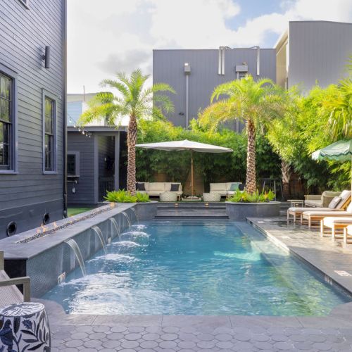 A modern pool with sun loungers, umbrellas, and palm trees, next to a grey building and surrounded by foliage.