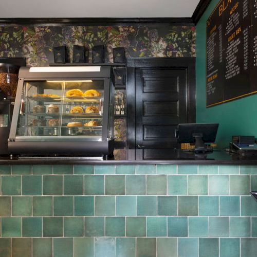 A café counter with a display case of pastries, a menu board, and a cash register, all set against green tiles and dark decor.