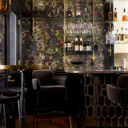 The image shows a cozy bar with patterned wallpaper, shelves of bottles, elegant bar stools, and warm lighting fixtures.