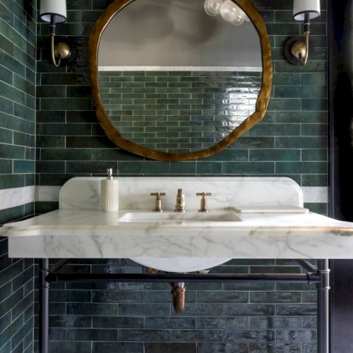 A bathroom with green tiles, a round mirror, two wall sconces, and a marble sink with gold fixtures, creating a stylish atmosphere.