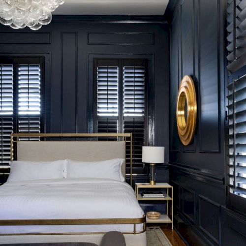 A bedroom with dark walls, a bed with white bedding, shuttered windows, a gold wall accent, and a lamp on a bedside table.