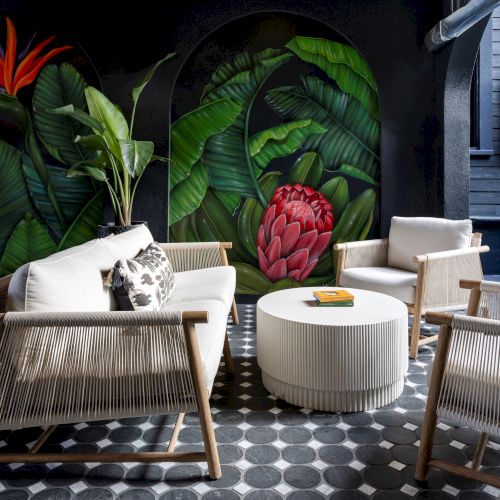 A stylish outdoor seating area with white furniture, a round table, and a vibrant tropical mural featuring large green leaves and red flowers.