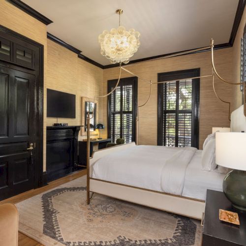 A cozy bedroom with a double bed, modern decor, black accents, a chandelier, and a small desk near a window with shutters.