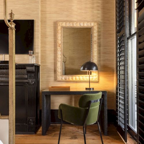The image shows a stylish room with a black fireplace, a wall-mounted TV, a desk with a lamp and mirror, a green chair, and shuttered windows.