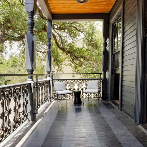 A charming porch with ornate black railings, a wooden ceiling, and two white chairs facing a small table, surrounded by trees.