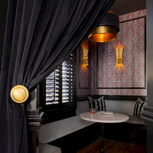 A cozy, elegant nook with a round table, cushioned seating, dark drapes, ambient lighting, and patterned wallpaper.