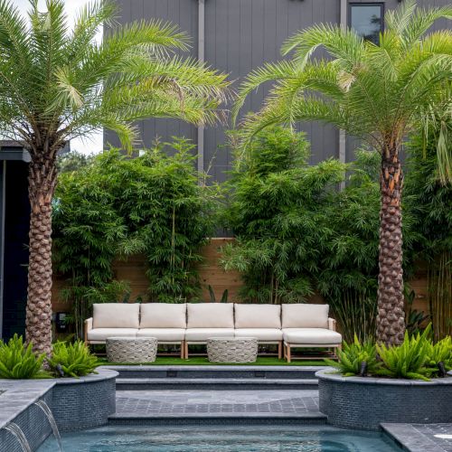 A backyard pool with two palm trees, a set of sofas, and lush greenery in the background creates a tropical oasis vibe.