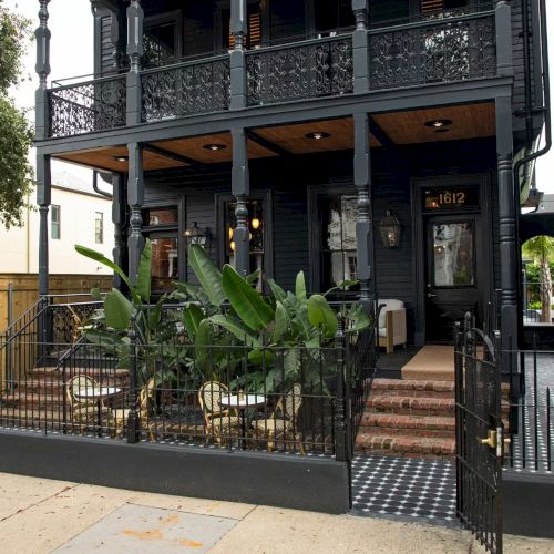 A charming, dark-hued building features a decorative wrought iron fence and a cozy outdoor patio with lush plants and seating.
