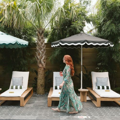 A woman in a floral dress walks by a pool with lounge chairs, umbrellas, and palm trees in the background.