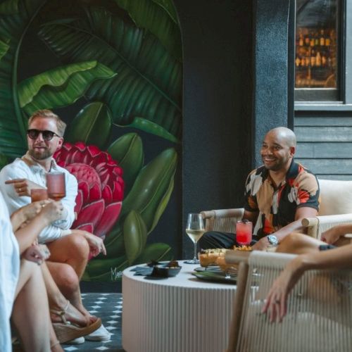 A group of people sit around a table with drinks, enjoying a casual gathering in a stylish setting with a vibrant mural background.
