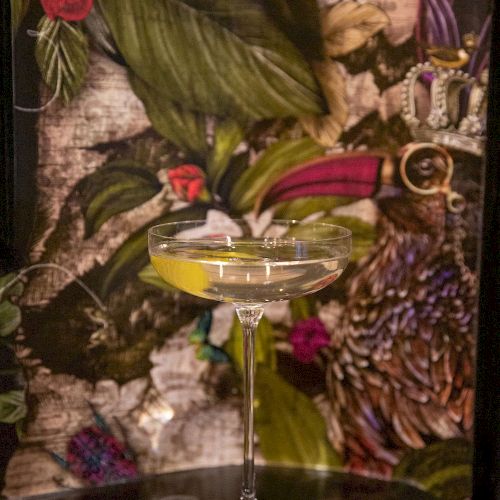 A cocktail in a glass with a lemon twist sits on a table against a colorful floral backdrop.