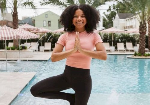 A person is doing a yoga pose by the pool, balancing on one leg with hands in prayer position, surrounded by palm trees and umbrellas.