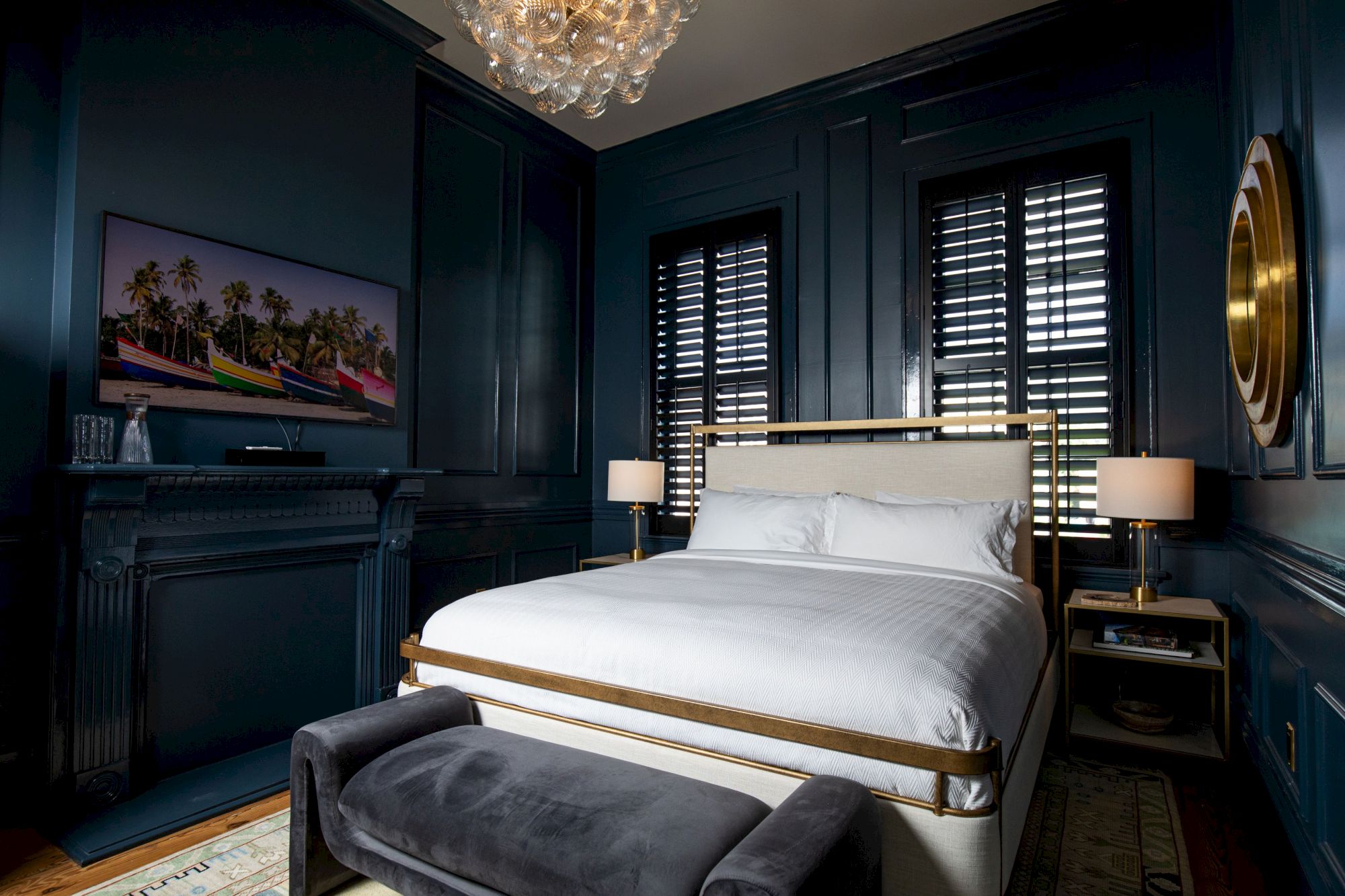 A cozy bedroom features a large bed, dark paneled walls, a chandelier, a wall-mounted TV, and two lamps on nightstands, all elegantly styled.