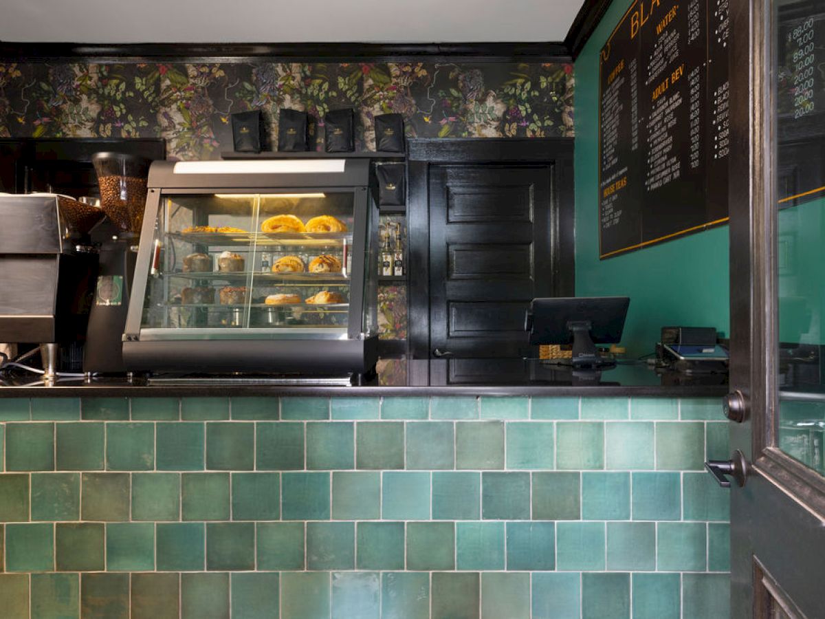 The image shows a cafe counter with a pastry display case, till, and menu board inside a shop with green tiled walls.