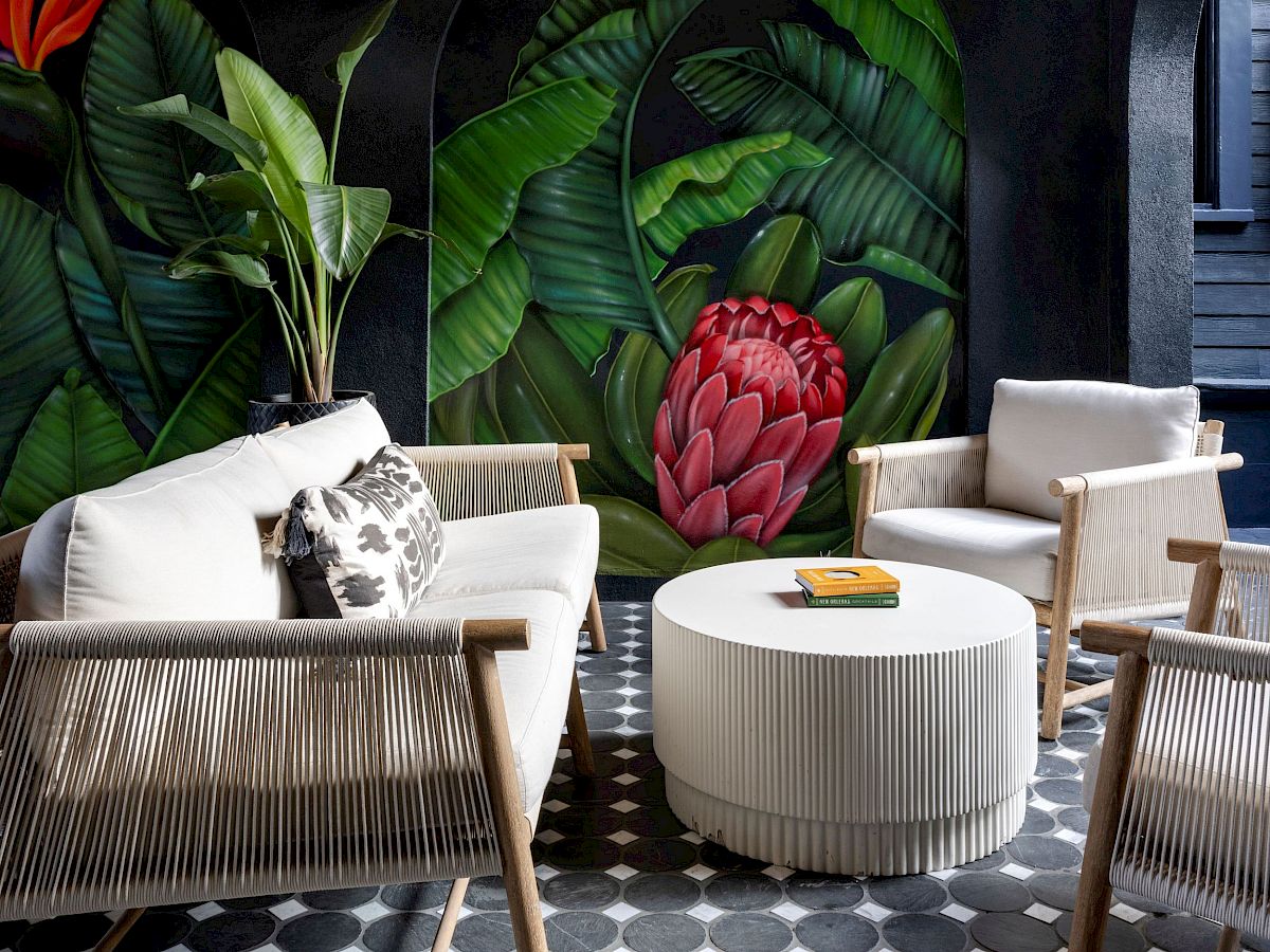 Outdoor seating area with white chairs, a round table, and tropical mural on the wall. Geometric floor tiles enhance the decor.