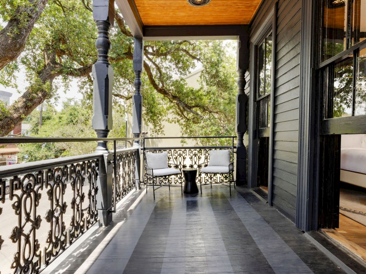 A wooden porch with ornate railing and two white chairs under a tree, providing a cozy outdoor seating area.