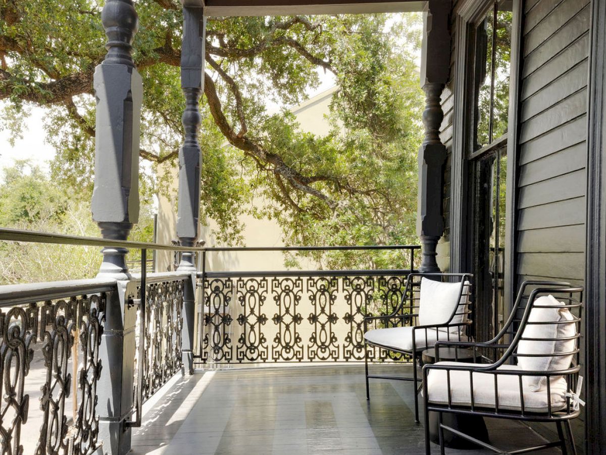A covered porch with decorative metal railing, two chairs, and a wooden ceiling, surrounded by trees for a serene outdoor setting.