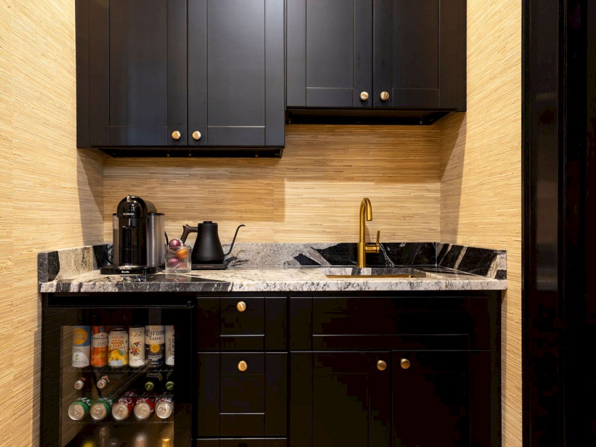 A compact kitchen area with black cabinets, a marble countertop, coffee maker, kettle, sink, and a mini fridge stocked with drinks.