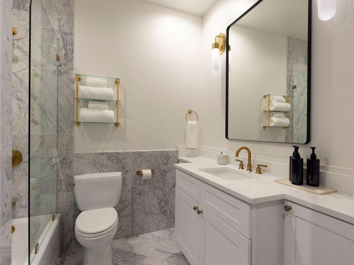 Elegant bathroom with marble tiles, gold accents, large mirror, and towels by the toilet and sink.