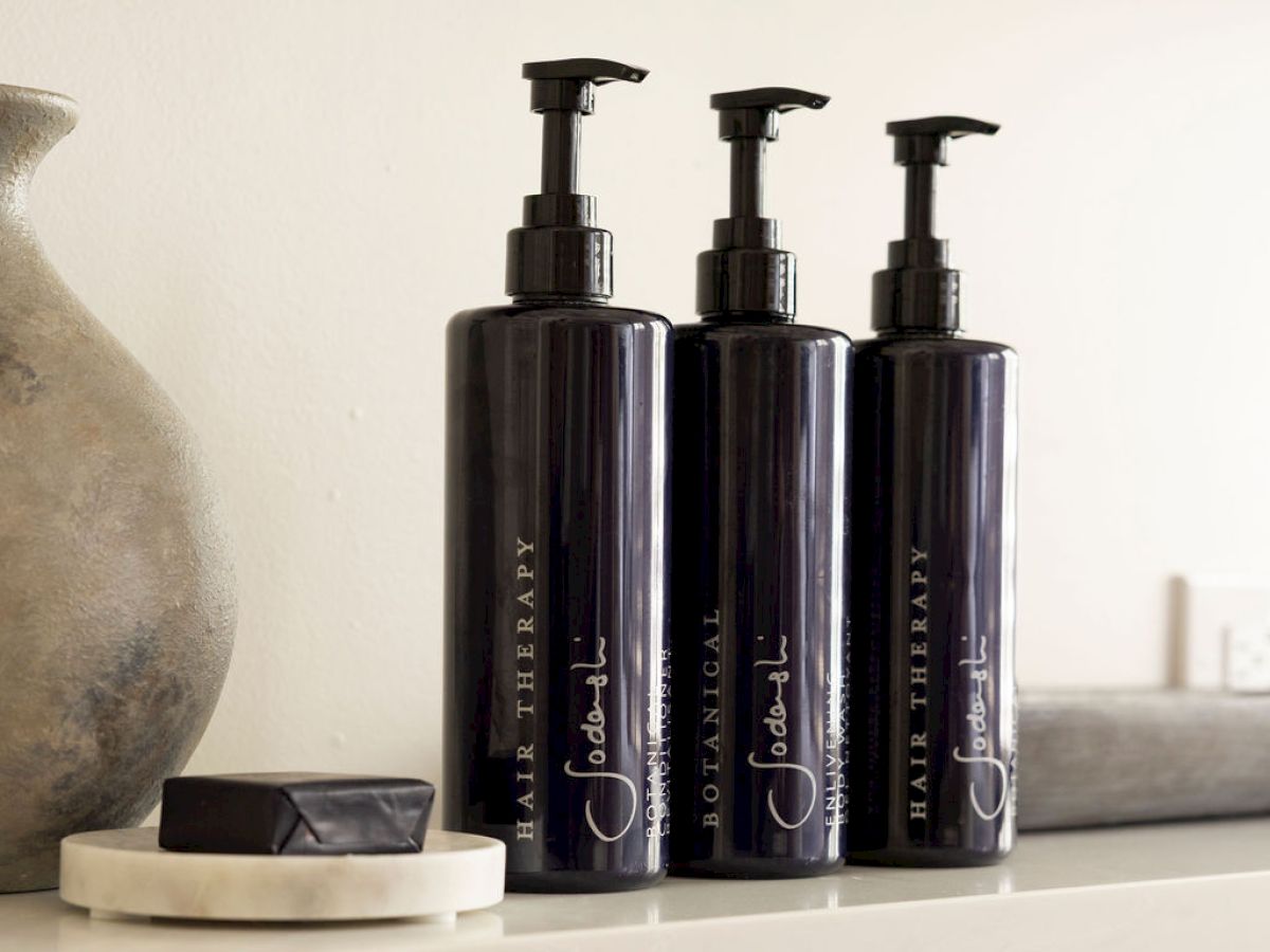 Three black pump bottles labeled "Botanical Hair Therapy" are on a shelf, alongside a decorative vase and a dish with a black soap.