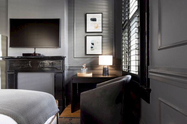 A stylish room with a wall-mounted TV, framed art, modern lamp, armchair, and side table by shuttered window, all in a sleek, dark theme.