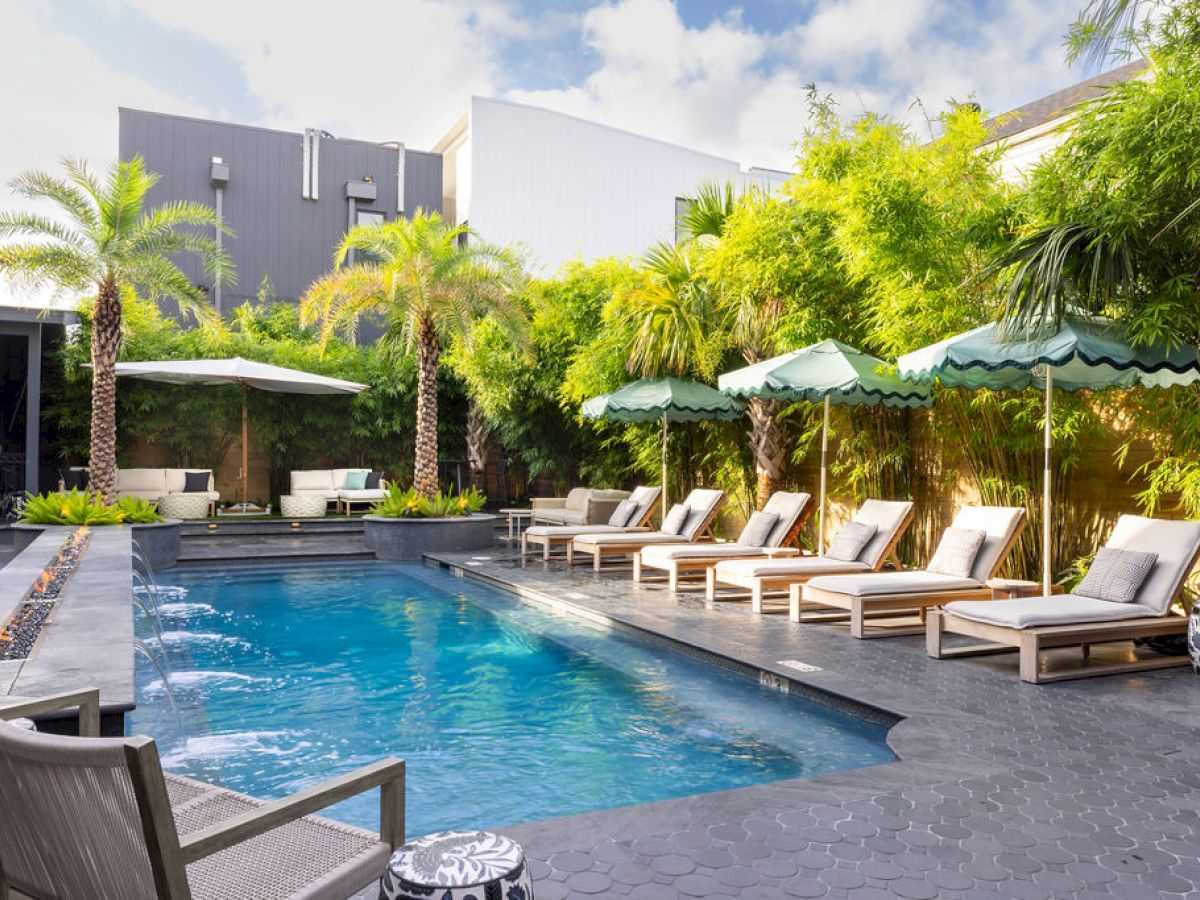 A modern poolside setting with lounge chairs, umbrellas, palm trees, and lush greenery in the background under a partly cloudy sky.