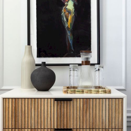 A wooden console with vases and a tray of glasses sits below a framed abstract painting in a minimalist interior setting.