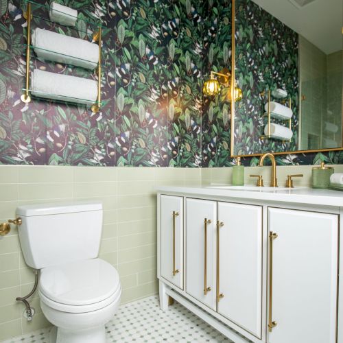 A bathroom with a leafy wallpaper, white vanity with gold fixtures, toilet, and patterned floor tiles, under warm lighting.
