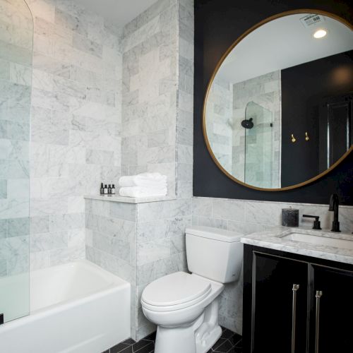 The image shows a modern bathroom with a marble-tiled wall, a bathtub, a toilet, and a vanity with a round mirror.
