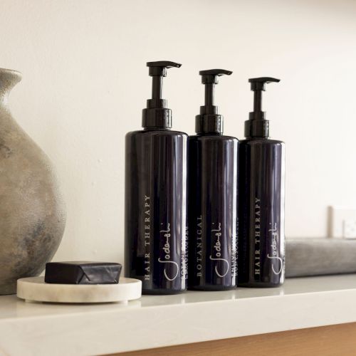 Three black pump bottles labeled for hair therapy and botanical use are on a shelf near a gray vase and a black rectangular object.