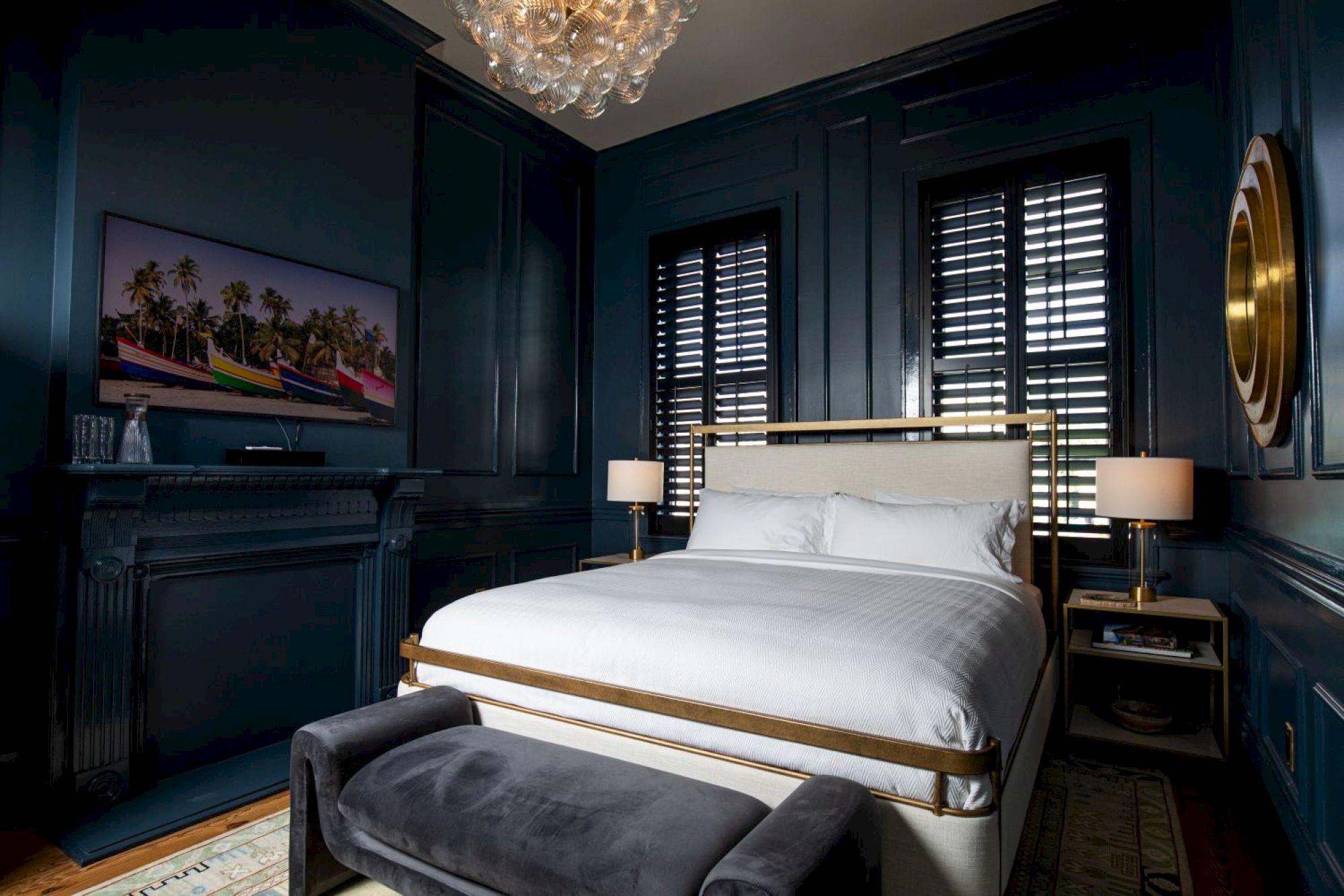 A cozy bedroom features a bed, dark walls, a chandelier, artwork, two lamps, and a bench, creating a warm and inviting atmosphere.