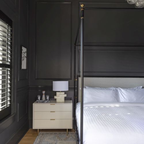 A stylish bedroom with a four-poster bed, gray walls, a white nightstand with a lamp, and a modern chandelier above.